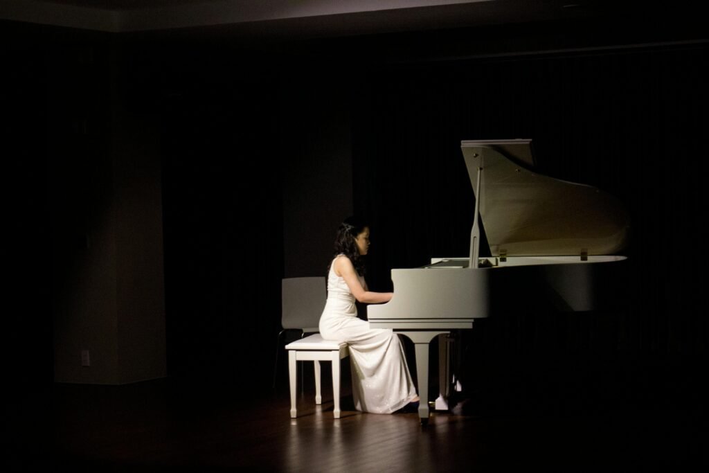 A woman in a white dress plays a grand piano under stage lights, creating a dramatic performance ambiance.